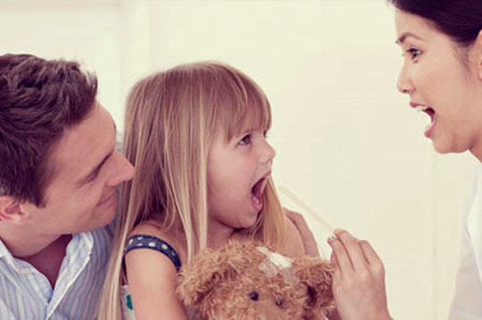 Mujer médico revisando la boca de una niña con una paleta mientras el padre está detrás